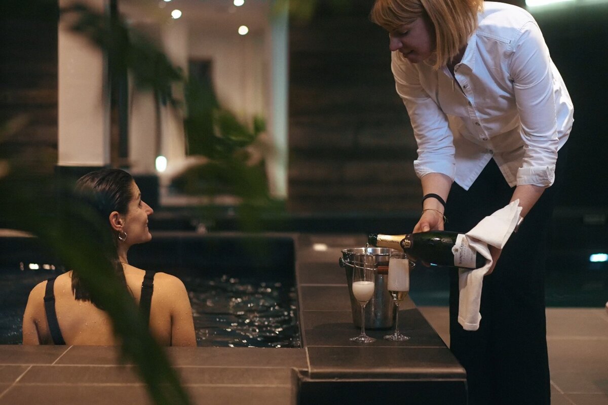 A woman pouring champagne into a bucket next to a woman in a bathtub