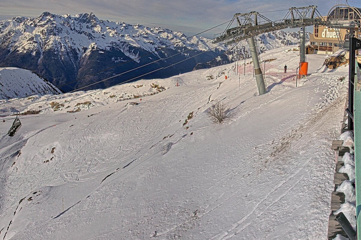 A snowy mountain ski resort scene featuring a ski lift, a building, and distant snow-capped peaks. Ski tracks are visible on the piste.