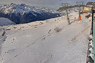 A snowy mountain ski resort scene featuring a ski lift, a building, and distant snow-capped peaks. Ski tracks are visible on the piste.
