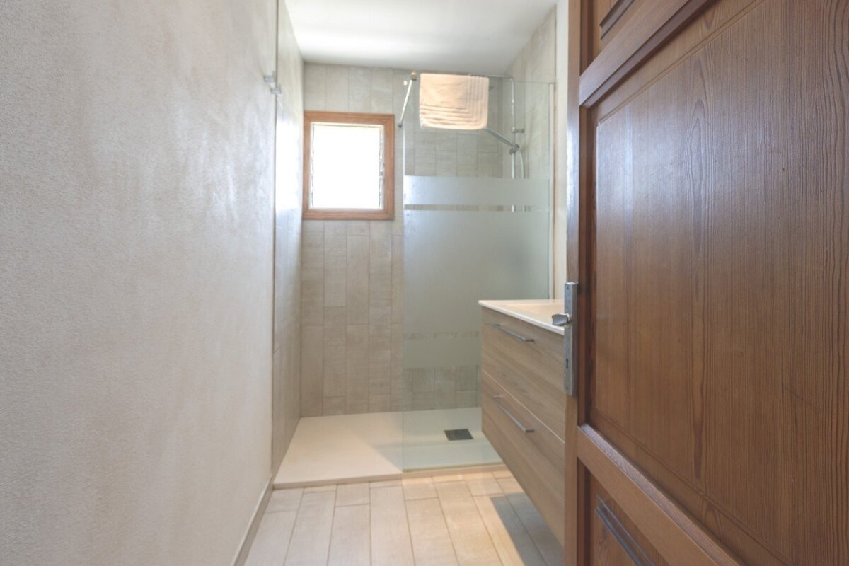 Bright, narrow bathroom with a walk-in shower (frosted glass), window, wooden vanity, and light tiled floor. An open wooden door frames the right.