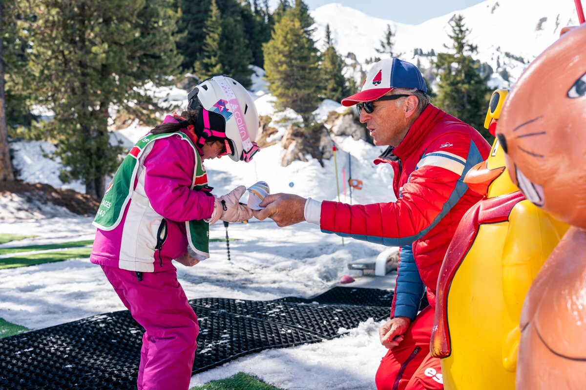A ski instructor helps a young girl in pink ski gear, exchanging a small toy on a snowy mountain slope with pine trees.