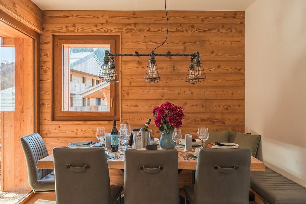 Cozy dining room with wooden walls, a set table, unique industrial pipe lighting, and a window view of chalet buildings.