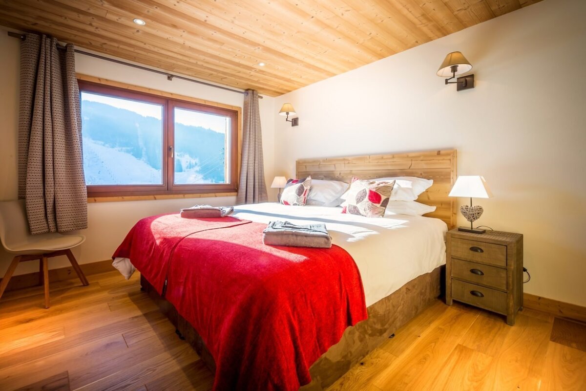 A cozy bedroom with a large bed, red blanket, wooden accents, and a window overlooking snowy mountains, creating a warm, inviting ski chalet feel.