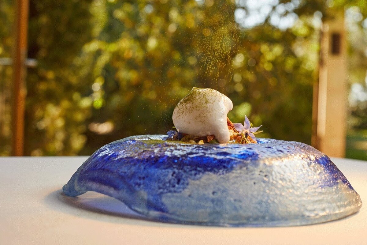 A delicate white foam dessert with green powder, flowers, on a textured blue dish, with golden dust falling and a blurred green background.