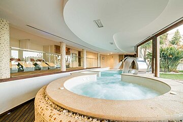 A modern indoor spa area featuring bubbling jacuzzi pools with water features, a glass wall overlooking lush greenery, and a relaxation zone with loungers.