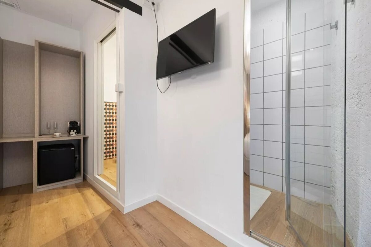 Modern room with wooden floor, mini-fridge, coffee maker, wall-mounted TV, and a glass shower stall with white tiles.