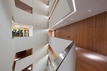 Sleek, multi-level modern interior featuring white walls, warm wood panels, strip lighting, and a glass-enclosed room on an upper floor.
