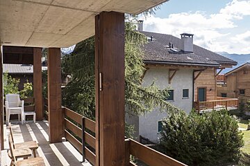 A wooden balcony with concrete ceiling overlooks houses, lush trees, and mountains under a partly cloudy sky.
