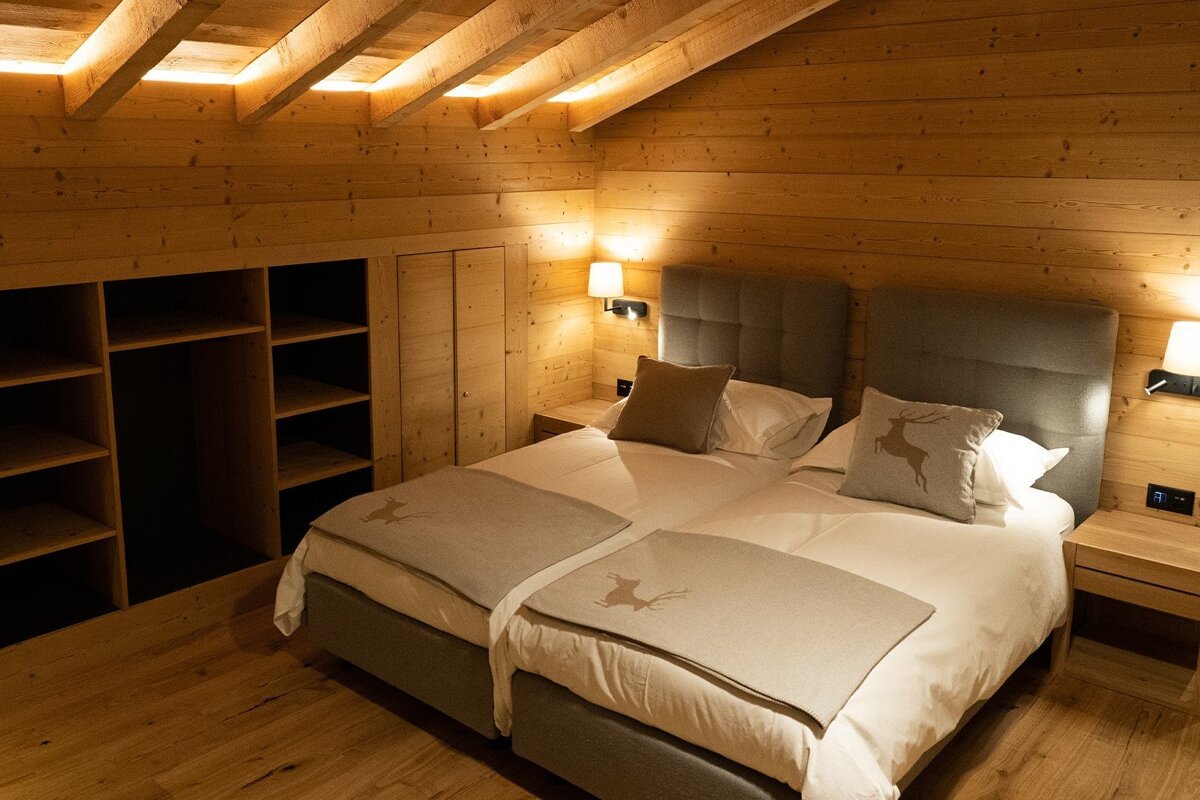 A cozy, wood-paneled bedroom features two beds with deer-themed bedding, bedside lamps, and open shelves. A slanted ceiling has ambient strip lighting.