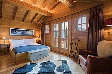Cozy wood-paneled chalet bedroom with a bed, cowhide rug, leather armchair, and French doors opening to a scenic mountain view.