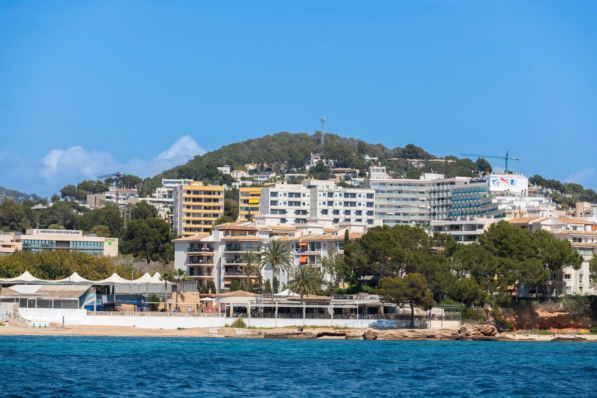 A picturesque coastal town featuring numerous buildings, green hills, a sandy beach, and clear blue water under a bright, sunny sky.