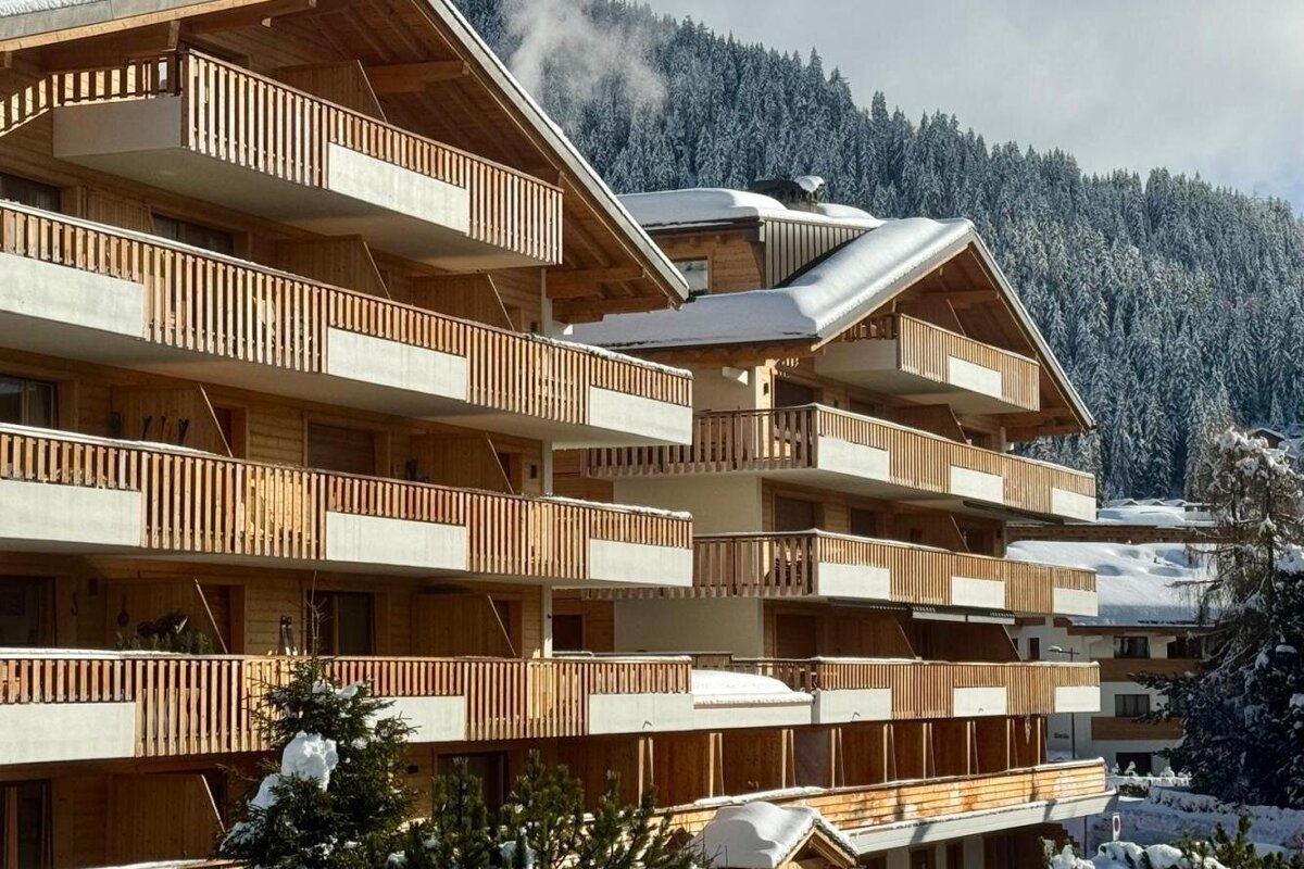 Snow-dusted wooden chalets with multiple balconies, set against a backdrop of snow-covered pine trees and mountains.