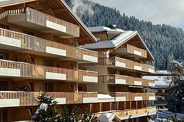 Snow-dusted wooden chalets with multiple balconies, set against a backdrop of snow-covered pine trees and mountains.