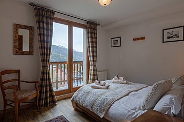 A cozy bedroom with a bed, wooden floor, and plaid curtains opens to a balcony with scenic mountain views.