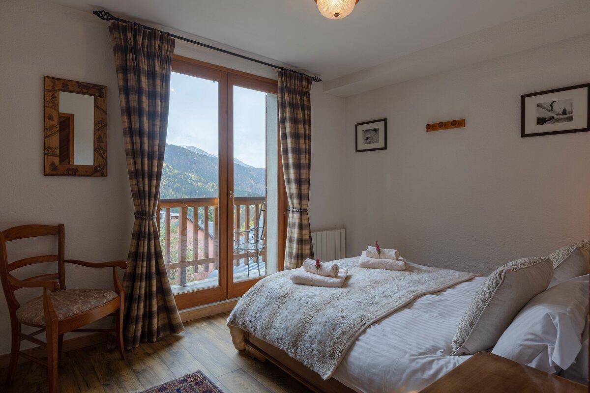 A cozy bedroom featuring a large bed, plaid curtains, and a balcony offering a stunning mountain view. White walls and framed pictures complete the decor.