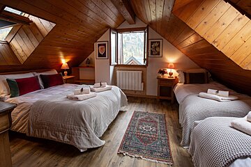 A cozy attic bedroom with warm wooden paneling, a double bed, and a single bed. A window reveals an outdoor view, illuminated by bedside lamps.