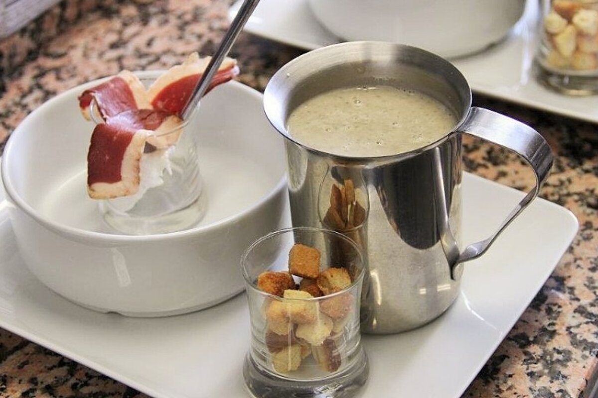A bowl of soup next to a pitcher of soup and a cup of croutons