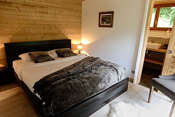 A cozy bedroom featuring a wooden accent wall, a large bed with a dark faux fur throw, warm lighting, and an ensuite bathroom with a sink.
