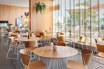 Bright, airy cafe interior with light wood tables and chairs, large window with blinds, plants, and minimalist decor.