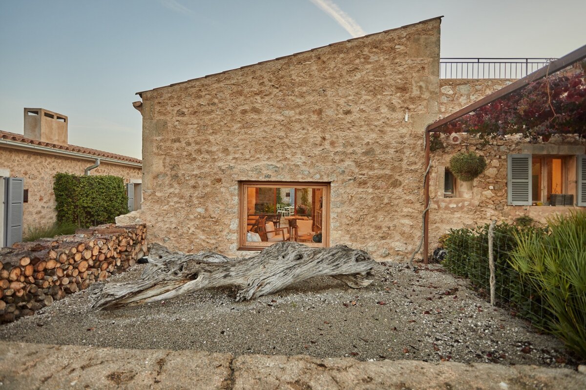 A stone building with a warm interior seen through a window, a large weathered log, and a stacked woodpile on gravel.