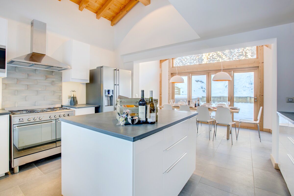 A bright, modern kitchen and dining room with a central island. Large windows show a snowy landscape, complemented by a wooden vaulted ceiling.