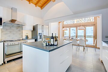 A bright, modern kitchen and dining room with a central island. Large windows show a snowy landscape, complemented by a wooden vaulted ceiling.