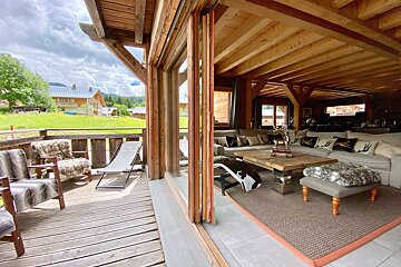 A rustic chalet living room opens to a wooden deck with fur chairs, offering views of green hills and houses under a cloudy sky.