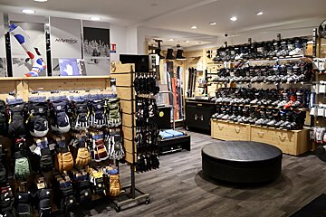 A well-lit ski shop showcasing rows of ski boots on shelves, various gloves on racks, and skis. A black ottoman rests on the dark wood floor.