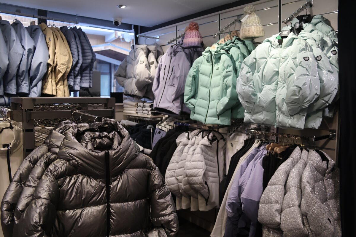 A clothing store displays racks of colorful winter jackets, including puffer coats in mint green, grey, silver, and black, alongside knit hats.