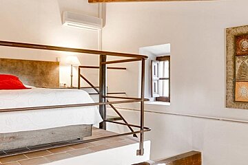 A bright mezzanine bedroom with a bed, red pillow, and metal railing. Features a small window, AC unit, and wall art on white walls.