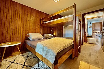 A cozy room features a wooden bunk bed, wood-paneled walls, and a patterned rug, with an open doorway leading to another room.