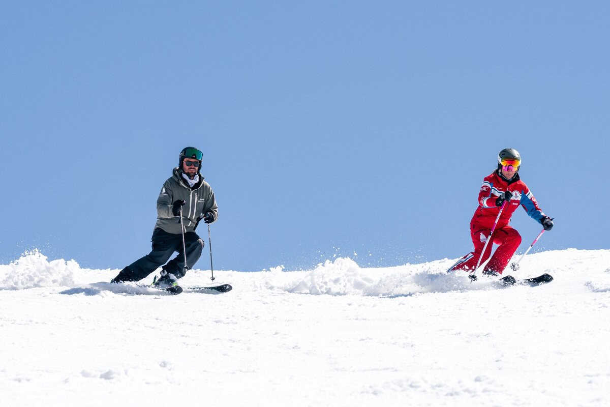Two skiers descend a sunny, snow-covered slope, kicking up powder against a bright blue sky.