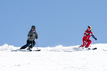 Two skiers carve down a sunny, snowy slope under a clear blue sky, one in green, the other in red, kicking up powder.