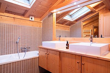 A cozy, wood-paneled bathroom featuring a brown tiled bathtub, dual rectangular white sinks on a wooden vanity, and a skylight.