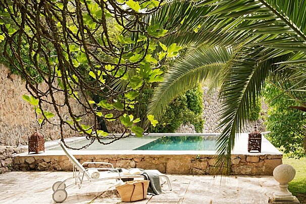 A relaxing outdoor pool area with a sunbed, basket, and lanterns, under shady fig and palm trees, with rustic stone walls.