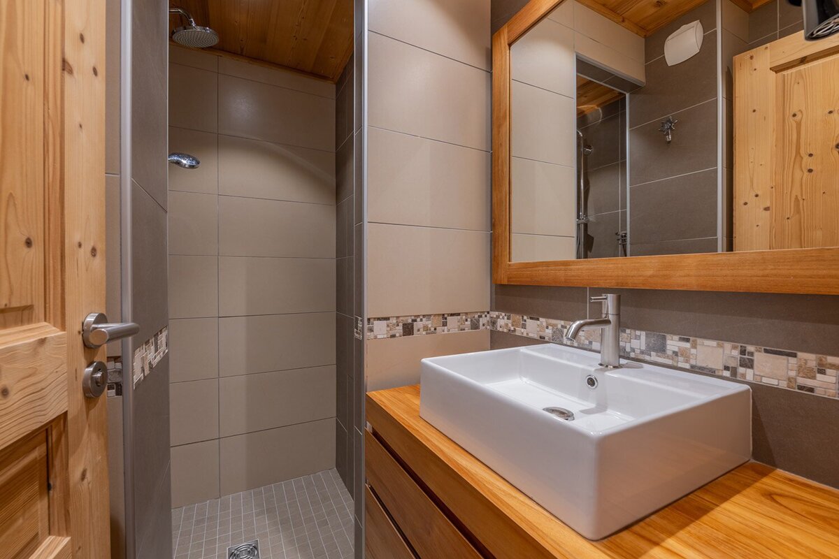 A modern rustic bathroom featuring a wooden vanity with a vessel sink, a walk-in shower, and natural wood accents on the door and ceiling.