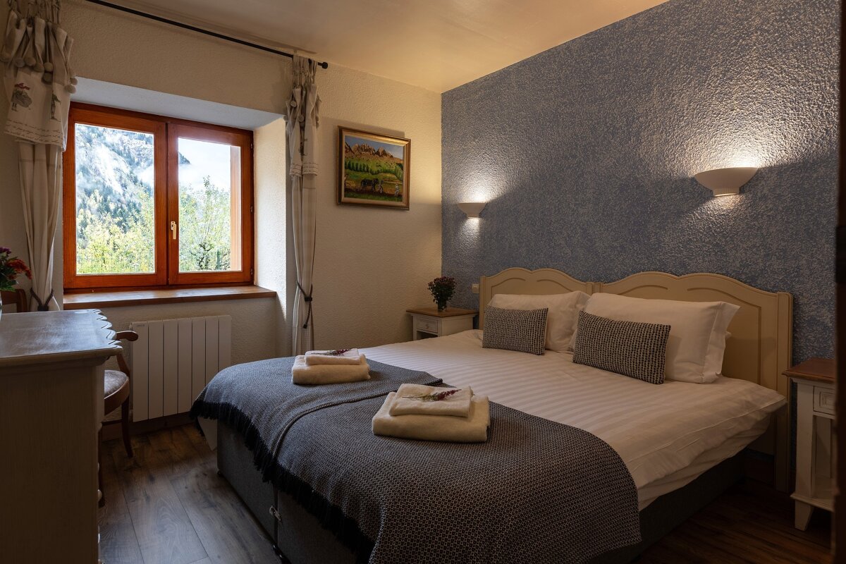 A cozy bedroom with a large bed, textured blue accent wall, and a wooden window framing a lush mountain view.