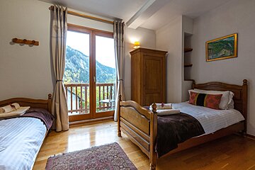 A cozy bedroom with two single beds, wooden furniture, and a balcony showcasing a beautiful mountain view. Hardwood floors and a patterned rug complete the space.
