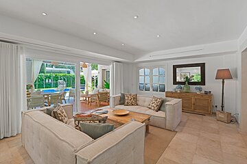 A bright, modern living room with cream sofas and a wooden coffee table, opening to a patio with outdoor seating and a swimming pool.