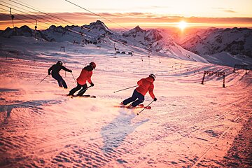 Three skiers descend a snowy mountain slope at sunset, bathed in warm golden light. Majestic peaks and ski lifts frame the vibrant winter scene.