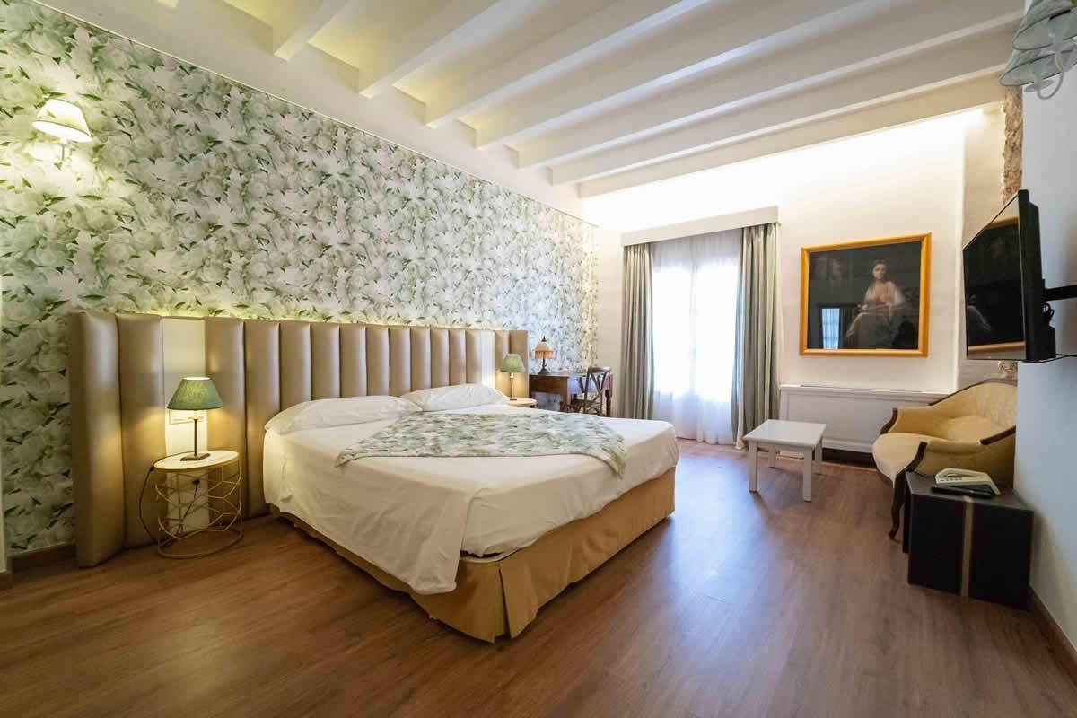 A modern hotel room with a large bed, padded beige headboard, green leafy wallpaper, wood floor, beamed ceiling, desk, and TV.