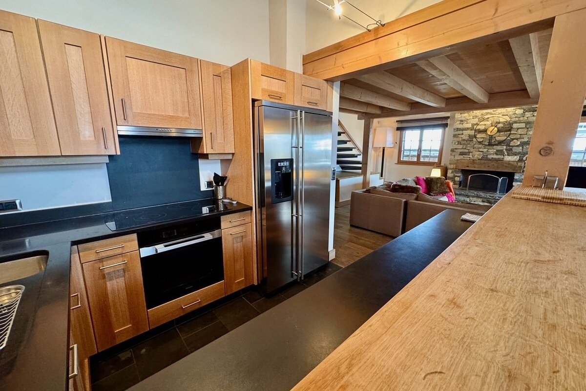 A modern kitchen with wood cabinets, black counters, & stainless steel appliances, opening to a living room with a stone fireplace & wood beams.