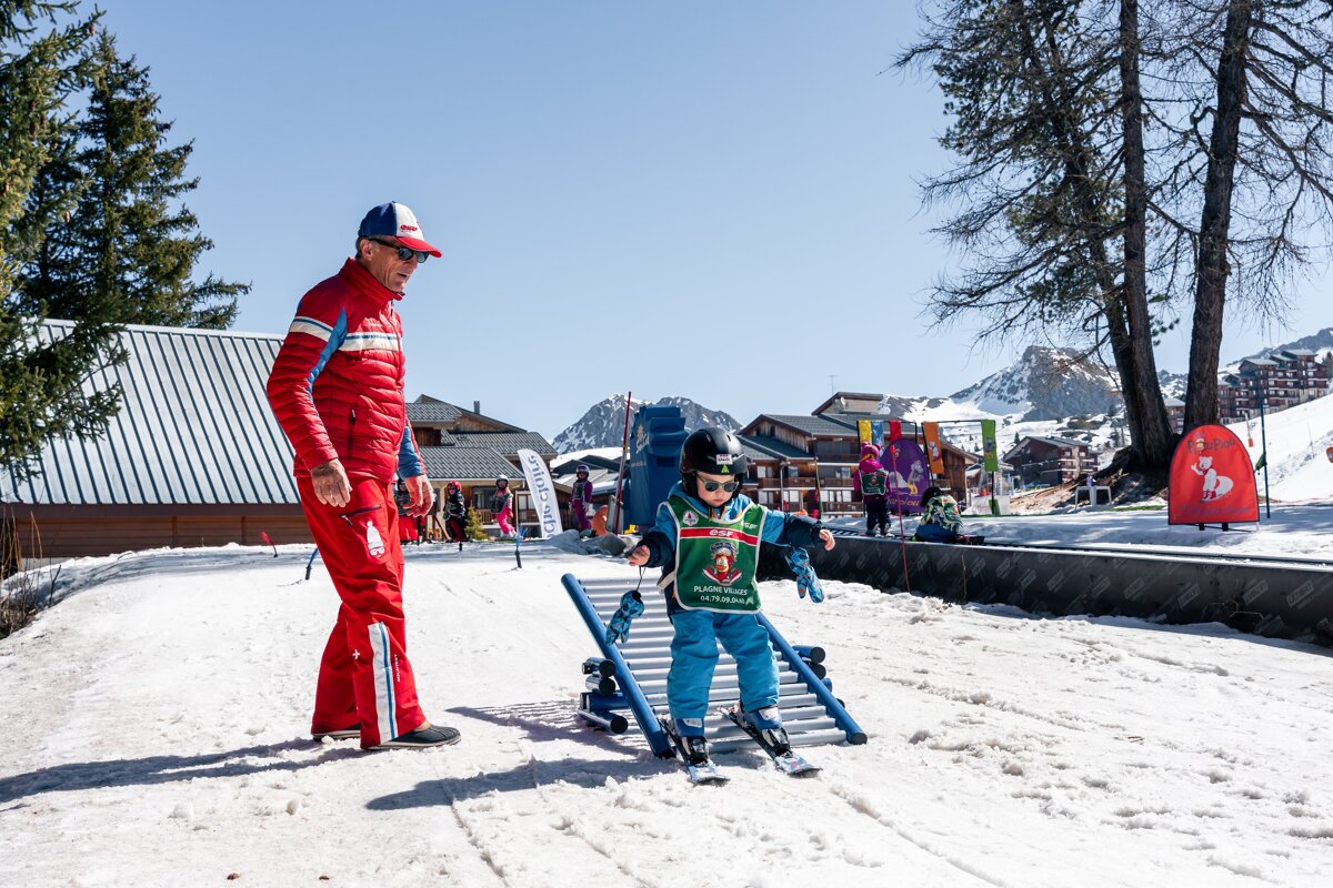 An adult instructor watches a child learning to ski on a training frame in a sunny, snowy mountain resort.