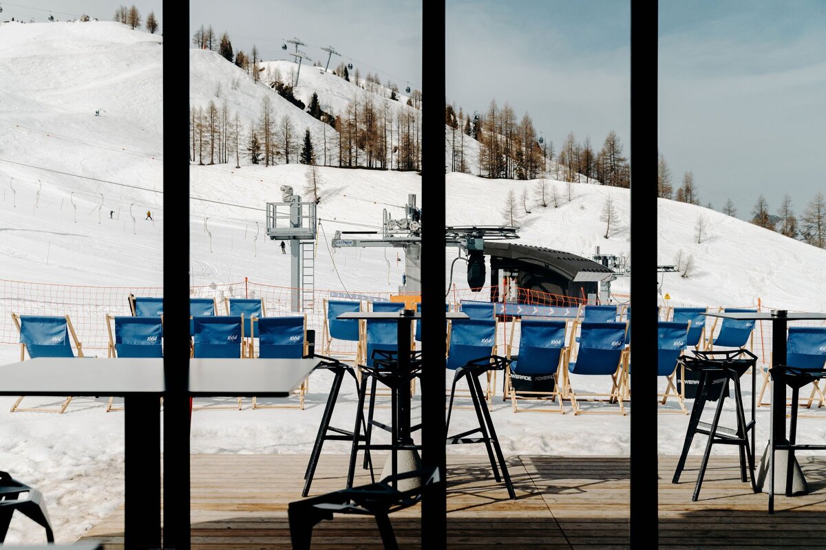 A window view of a snowy ski resort with mountains, ski lifts, and empty blue lounge chairs on a wooden deck, framed by dark vertical bars.