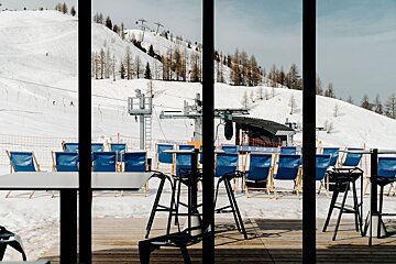 A window view of a snowy ski resort with mountains, ski lifts, and empty blue lounge chairs on a wooden deck, framed by dark vertical bars.