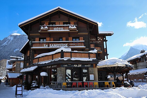 A multi-story, snow-covered wooden chalet hotel, 'Le Samoyède,' featuring 'Cafe Le 19.38,' stands amidst snowy mountains under a bright blue sky.