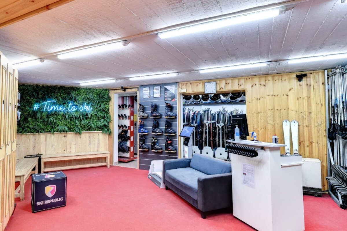 A brightly lit ski rental shop featuring ski boots, skis, benches, a sofa, and a green wall with a '#Time to ski' neon sign.