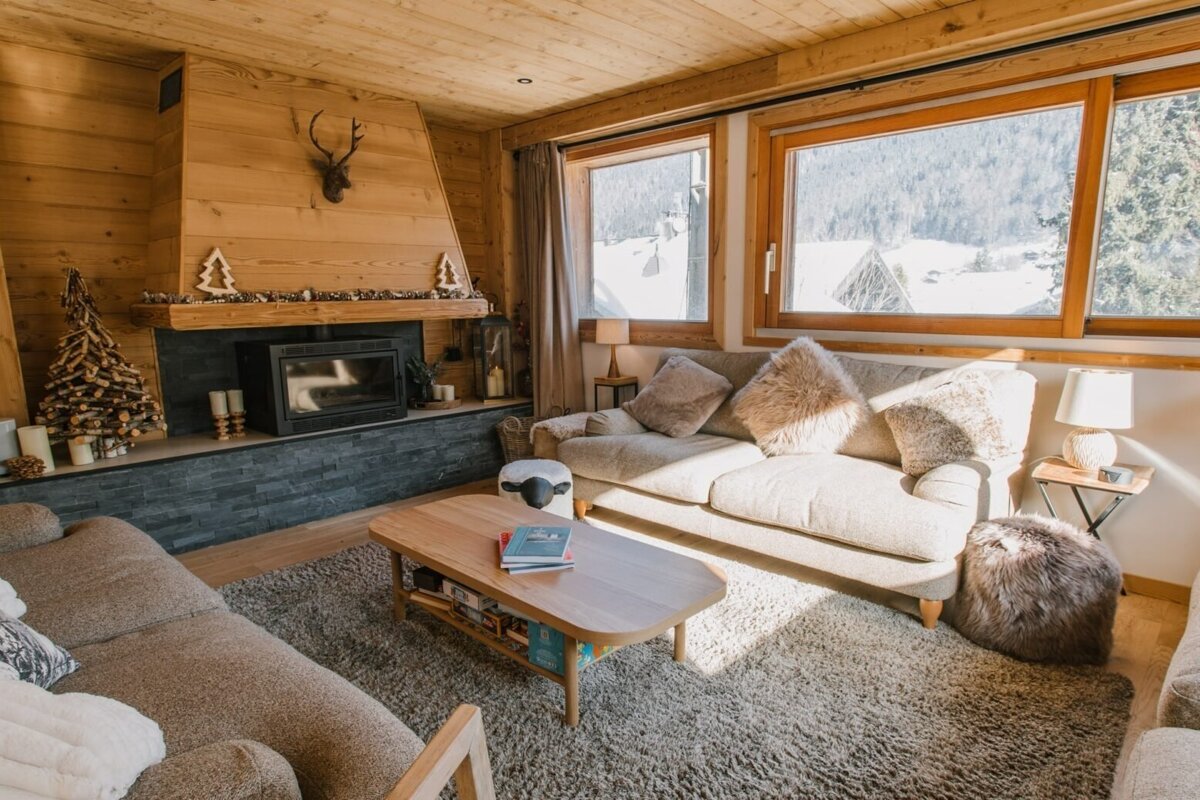 A cozy, wood-paneled ski chalet living room with a stone fireplace, comfy sofas, and large windows revealing a scenic, snowy mountain view.
