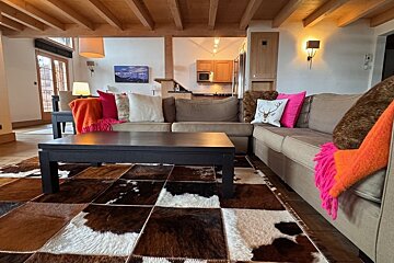 A cozy, chalet-style living room with a large L-shaped sofa, cowhide patchwork rug, wooden beams, and bright accent pillows/throws. Kitchen visible.
