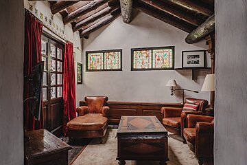 Inviting room with exposed wood beams, red drapes, stained glass, and leather chairs. A wooden chest serves as a coffee table.